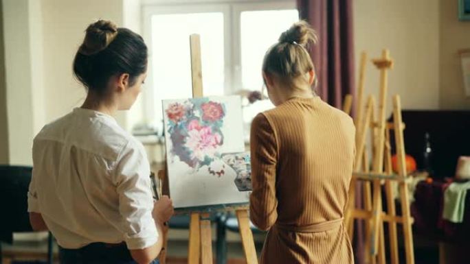 艺术系学生在画布上画花的背景图和她乐于助人的老师站在附近检查她的作品。视觉艺术与教育理念。