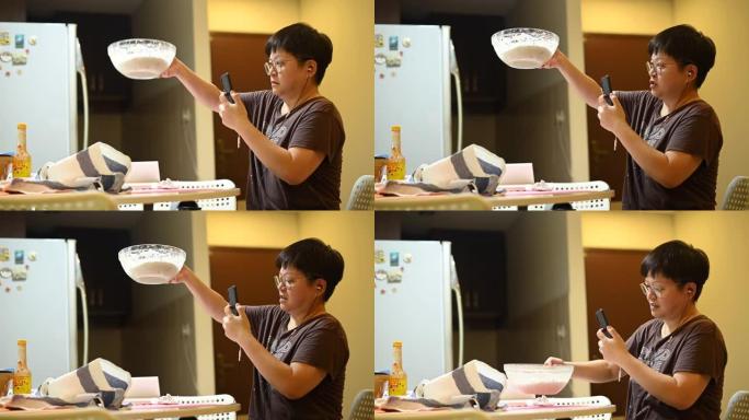 一位亚洲华人待在家里的母亲正在准备面团，同时使用电话在线检查食谱和在线指导课程