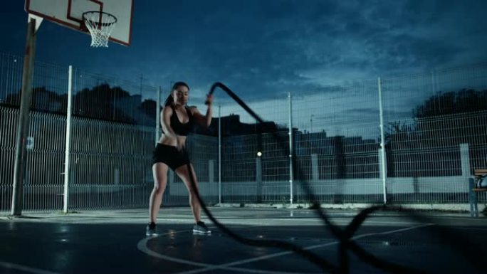 美丽精力充沛的健身女孩用战斗绳锻炼。她正在一个有围栏的室外篮球场里锻炼身体。居民区下雨后的晚间录像。
