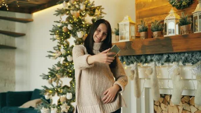 无忧无虑的女孩在圣诞节那天站在新年树和壁炉旁自拍，拿着智能手机摆姿势拍照。假期和照片概念。