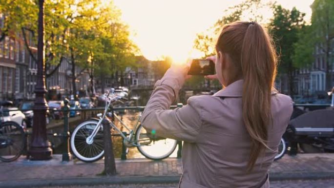 阿姆斯特丹度假的女性游客日落时站在运河上的桥上，用智能手机拍照
