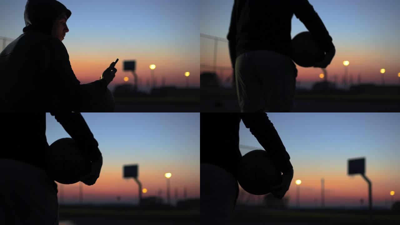 带着智能手机和篮球的年轻男子在黄昏时走出篮球场