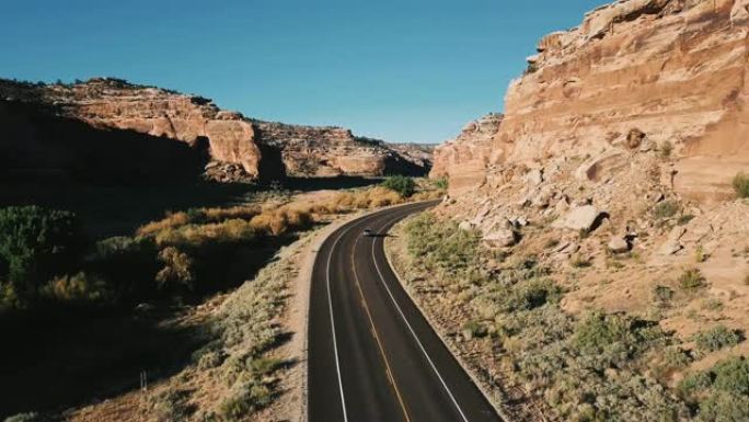 摄像机跟随汽车在覆盖着沙子和岩石的陡峭阳光峡谷山脉之间的高速公路上行驶。