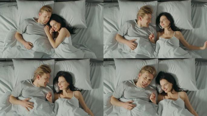 幸福的年轻夫妇在床上拥抱在一起。美丽的女孩和英俊的男孩在悠闲的早晨，在早晨慢慢醒来。自上而下。