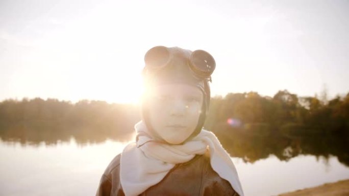 穿着复古飞行员服装的小飞行员男孩，戴着围巾和眼镜，看着相机在日落湖慢动作中微笑。