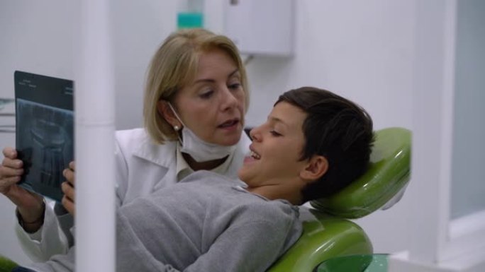 可爱的女牙医与儿科患者交谈，当他躺在牙医椅子上时，向他展示了牙齿的x射线