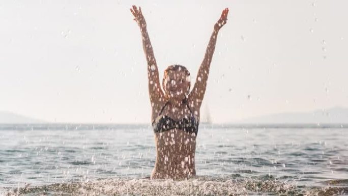 穿着比基尼的年轻幸福的女人在暑假期间站在海边的海洋里泼水
