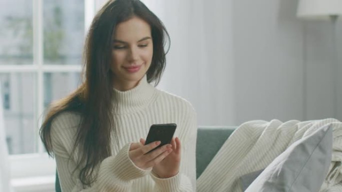 年轻女子使用智能手机，坐在椅子上喝茶。性感的女孩穿着毛衣上网，在社交媒体上发帖，在舒适的公寓里放松时
