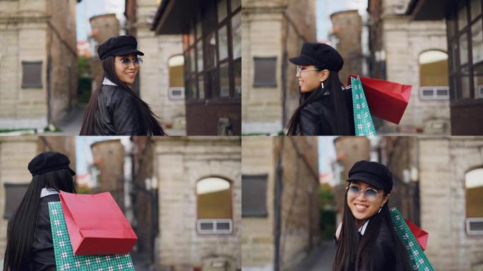 迷人的亚洲女孩拿着购物袋走在街上，转身，微笑着看着相机的慢动作肖像。人、消费主义和城市生活观。