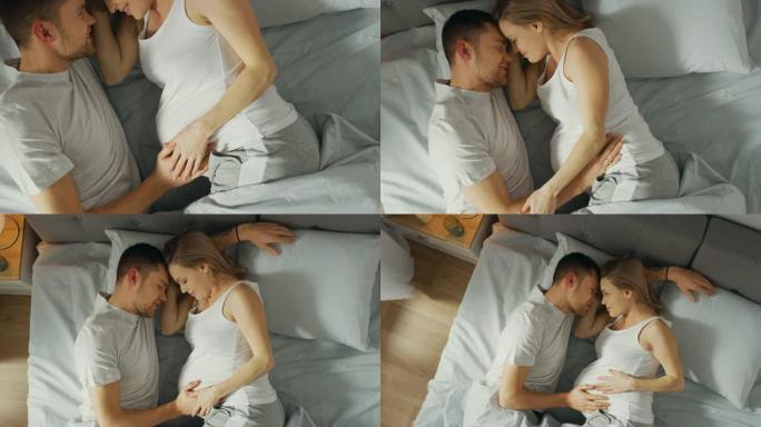 幸福的年轻夫妇在床上拥抱在一起，年轻的女人怀孕了，有爱心的伴侣温柔地抚摸着她的肚子。俯视图缩小相机拍