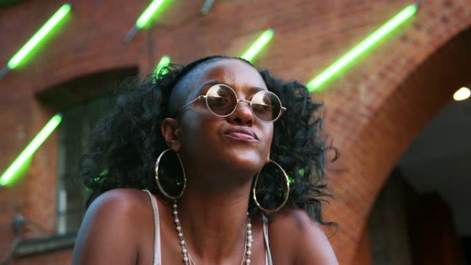 时尚年轻黑人女子戴着圆形太阳镜坐在街上，低角度，特写，背景中模糊的霓虹灯条