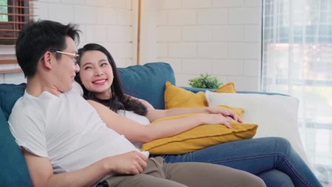 亚洲情侣在家里的客厅拥抱在一起，甜蜜的情侣在家放松时躺在沙发上享受爱情时刻。生活方式夫妇在家放松的概