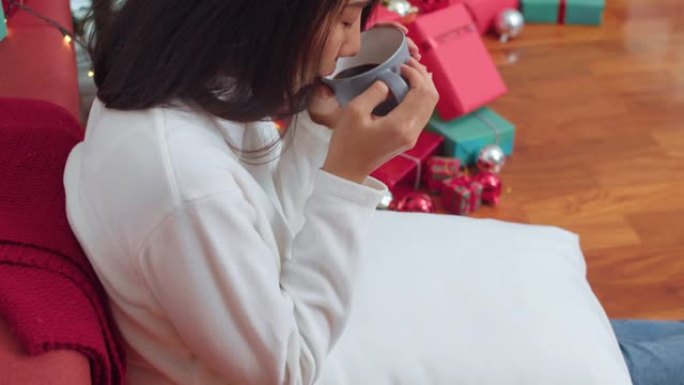 亚洲女性庆祝圣诞节。女性青少年放松快乐在沙发上的圣诞树附近喝咖啡在家里的客厅享受圣诞节的寒假。
