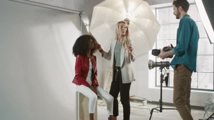 照片拍摄的后台: 化妆师在美丽的黑色模特上涂抹口红化妆，卷发茂盛。摄影师准备拍照。时尚杂志工作室摄影