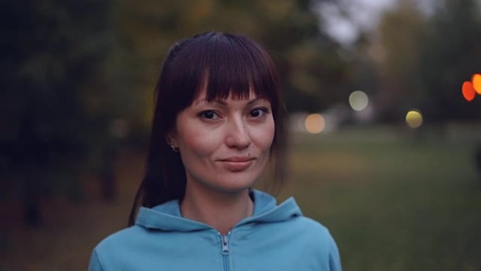 美丽的年轻女运动员的肖像，长长的黑发和棕色的眼睛看着相机，微笑着站在秋天的夜晚，穿着连帽衫站在户外。
