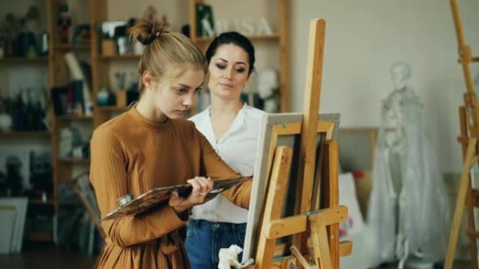 经验丰富的美术老师正在与漂亮女孩talanted学生一起在充满艺术品的工作室里在室内绘画和谈话分享经