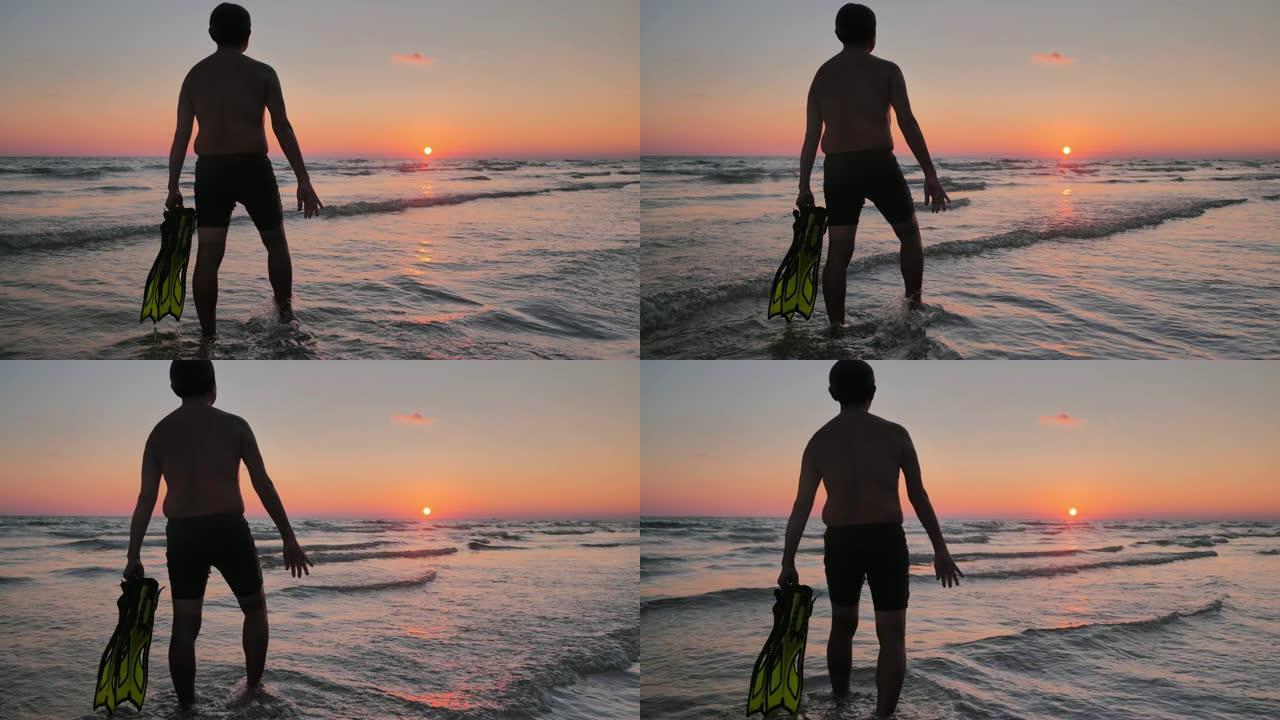 老人拿着面具和脚蹼在日落时在海上游泳的剪影。老人在暑假放松。真实的身体