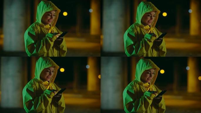 穿着黄色雨衣的美丽年轻女孩在雨中使用智能手机。收到通知时，她在微笑。天气多雨，现在是晚上。
