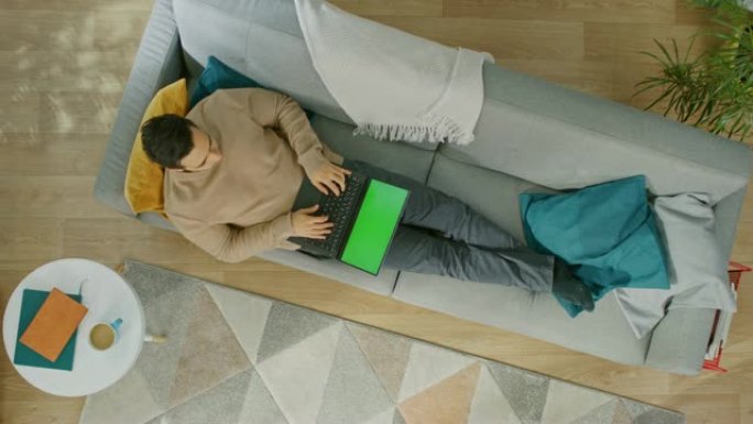 穿着棕色套头衫和灰色牛仔裤的年轻人躺在沙发上，使用带有绿色屏幕的笔记本电脑。舒适的客厅，配有现代室内