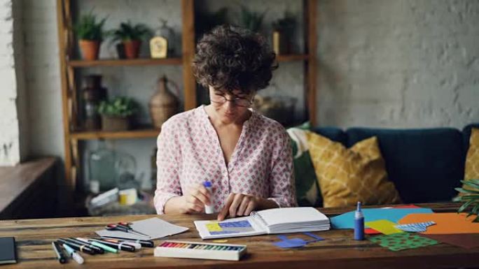 戴眼镜的年轻黑发女人在设计插图时，用胶条和剪刀在笔记本上剪下飞溅纸，然后将人物粘贴在笔记本上。艺术家