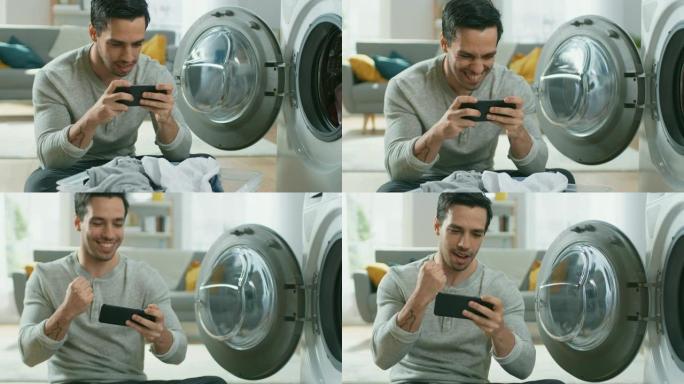 穿着灰色牛仔裤和外套的英俊微笑的年轻人坐在洗衣机前，在智能手机上玩游戏。他给洗衣机装上脏衣服。明亮宽