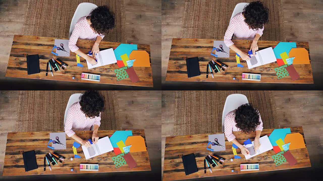 忙碌的女设计师在笔记本上使用胶水粘纸图的俯视图-独自在家工作。创意、设计和现代艺术理念。