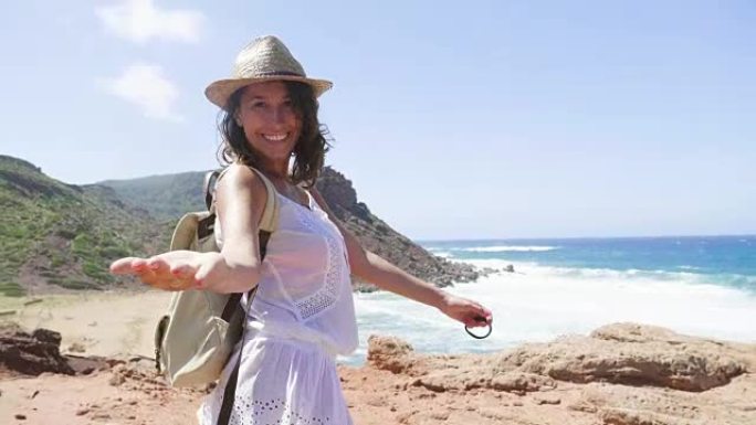 一位美丽的年轻游客 (女孩) 的肖像，穿着白色连衣裙，穿着背包，戴着草帽，沿着岩石，海洋背景和山脉行