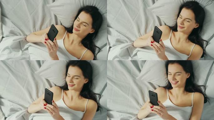 美丽的黑发肖像在床上使用触摸屏智能手机，与朋友聊天，使用社交媒体，发布图片，分享想法。自上而下的相机
