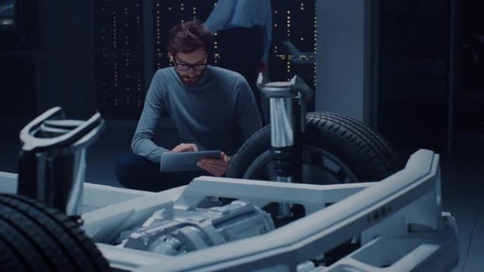 汽车设计工程师坐在混合动力电动汽车底盘平台原型旁边，使用平板电脑进行设计增强。带有悬架、车轮、发动机