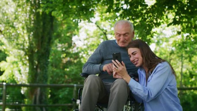 坐在轮椅上的快乐孙女和祖父的慢动作在阳光明媚的日子里，在绿色公园的手机上看他们的照片或在互联网上导航