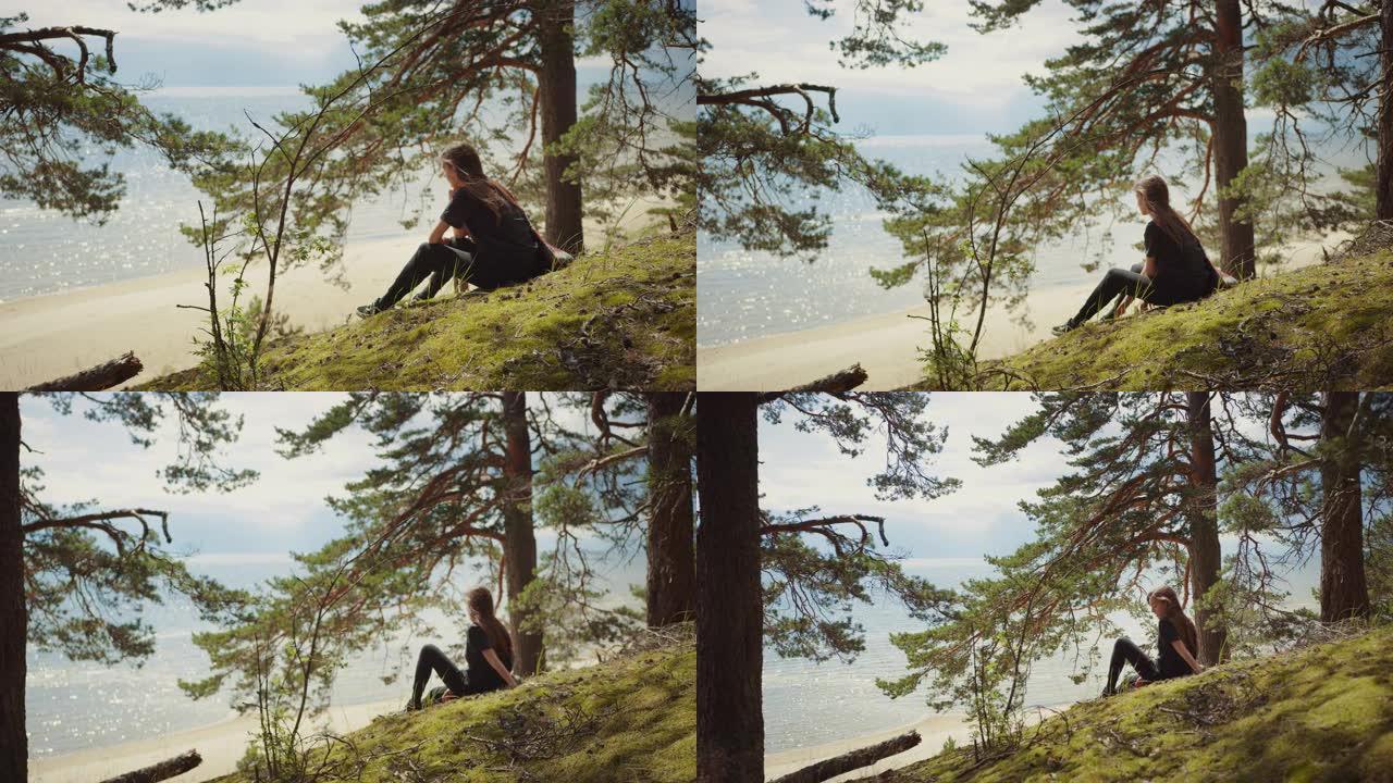 穿着黑色衣服的年轻美女坐在欧洲松树林中。这是大自然温暖的晴天。她白天梦想在边缘，看着海景。