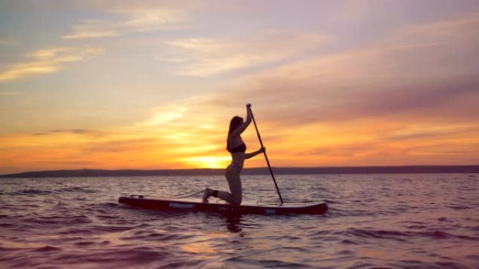 日落背景上的冲浪女孩，侧视图。女运动员在冲浪板上划着桨。
