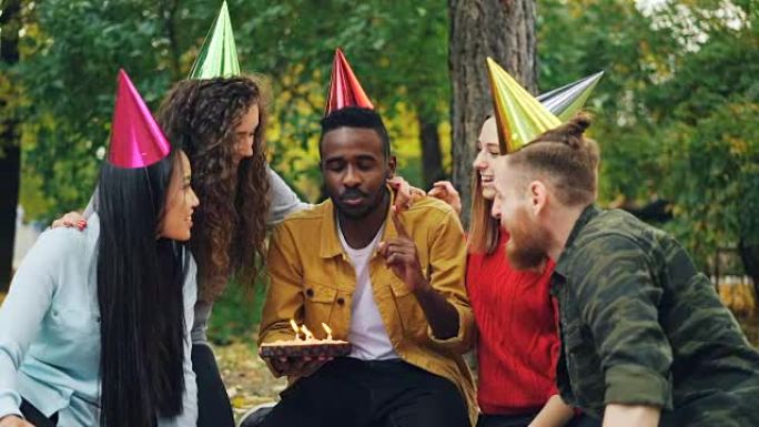 快乐的非洲裔美国人的慢动作许愿，然后在蛋糕上吹蜡烛，而他戴着派对帽子的朋友正在祝贺他。户外派对概念。