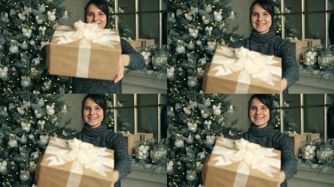 微笑的女孩拿着礼品盒的肖像，然后伸出双手，向圣诞节表示祝贺。装饰的枞树和壁炉架是可见的。