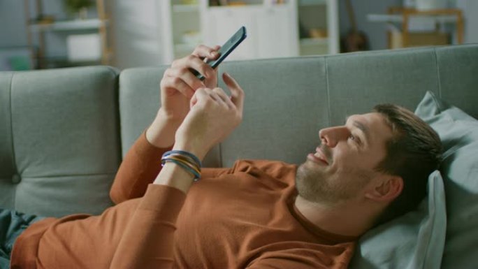 英俊的年轻人躺在家里的沙发上使用Smarphone，进行滑动和触摸手势。侧视图摄像机角度。