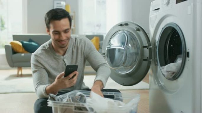 穿着灰色牛仔裤和外套的英俊微笑的年轻人坐在洗衣机前，使用智能手机。他给洗衣机装上脏衣服。明亮宽敞的客