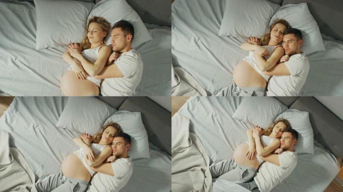 幸福的年轻夫妇一起睡在床上，可爱的年轻人在睡觉时互相抱着。美丽的女人怀孕了，有爱心的伴侣支持她。