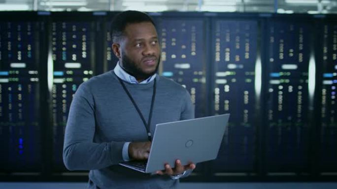 黑色数据中心it技术人员站在服务器机架走廊的中间，上面放着一台笔记本电脑。他正在目视检查工作服务器机