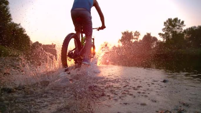 慢动作: 运动员在朝着日落方向的浅溪流中骑自行车。