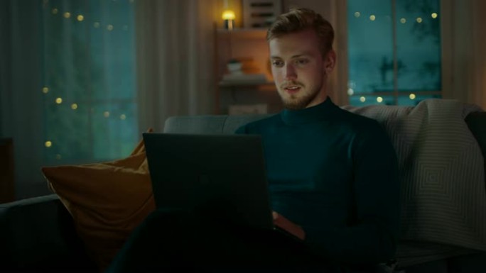 晚上，英俊的年轻人坐在客厅的沙发上，在笔记本电脑上工作。他使用计算机并通过互联网浏览，充满乐趣和微笑