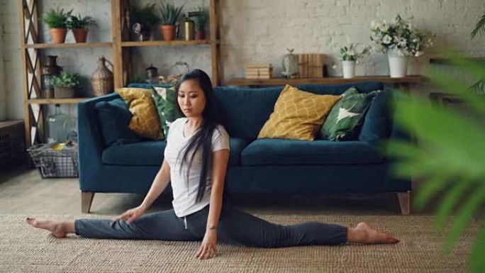 亚洲女体操运动员正在家里训练坐在客厅地毯上的劈腿。积极的生活方式，灵活性和运动的年轻人概念。
