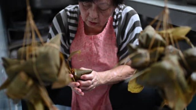 一位亚洲华裔高级妇女在厨房里准备zong子中国饺子，并将其包裹起来，为端午节做准备