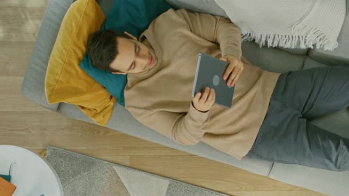 穿着棕色套头衫和灰色牛仔裤的年轻人躺在沙发上，用平板电脑。他快乐而微笑。舒适的客厅，配有现代室内植物