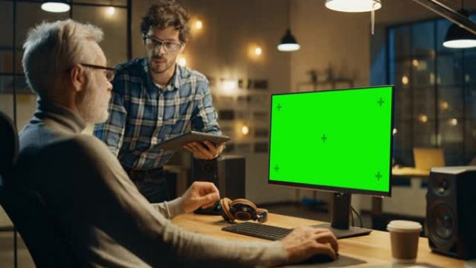 晚上，创意中年男子在台式计算机上工作，绿色模拟屏幕，与使用平板电脑的年轻专家交谈。时尚的大城市办公室
