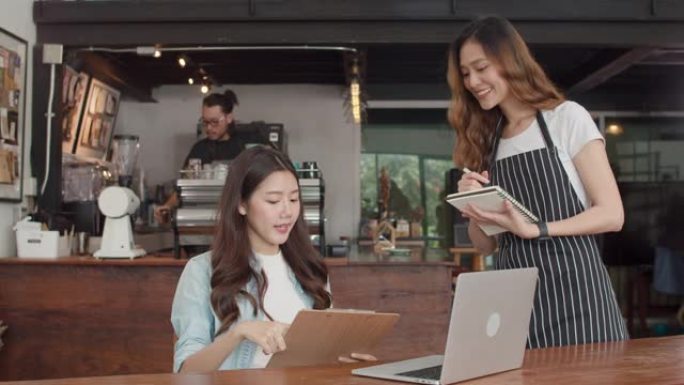 快乐的亚洲女服务员带着笔记本在城市咖啡馆接受年轻客户女孩的订单菜单。