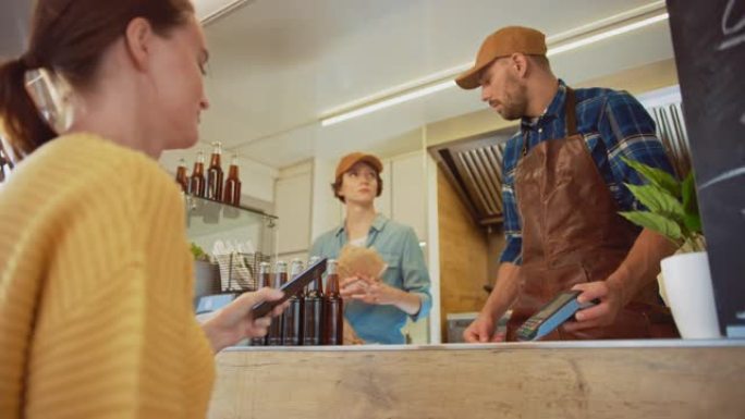 食品卡车员工将汉堡交给一个快乐的年轻女子。年轻女士正在智能手机上使用NFC非接触式支付来支付食物。街