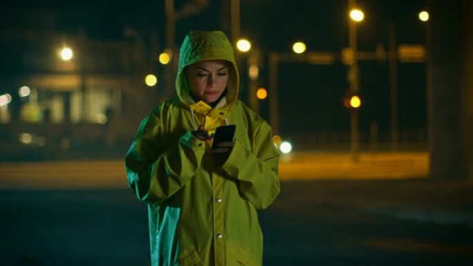 穿着黄色雨衣的美丽年轻女孩在雨中使用智能手机。收到通知时，她在微笑。天气多雨，现在是晚上。
