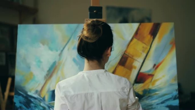 熟练的艺术家漂亮女人正在用丙烯酸颜料在画布上画海景海浪和船，拿着调色板和画笔，创造杰作。人和艺术概念