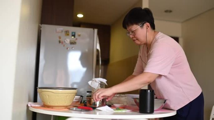 一位亚洲华人留在家里的母亲在使用虚拟助手智能扬声器制作食谱和指导时准备面团