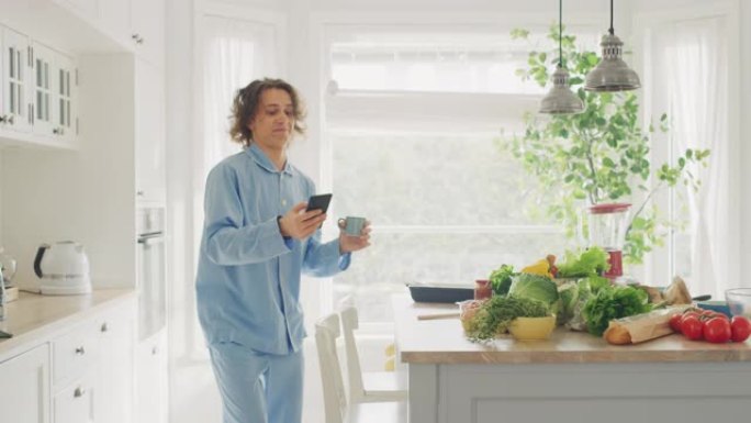 穿着蓝色睡衣，长发的快乐年轻人在家里跳舞。他正在手机上听音乐。精力充沛的人在厨房里使用智能手机，里面